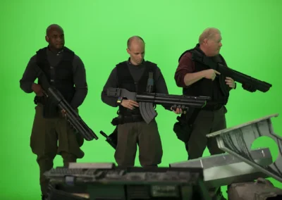 3 actors on a green screen holding large laser rifles.